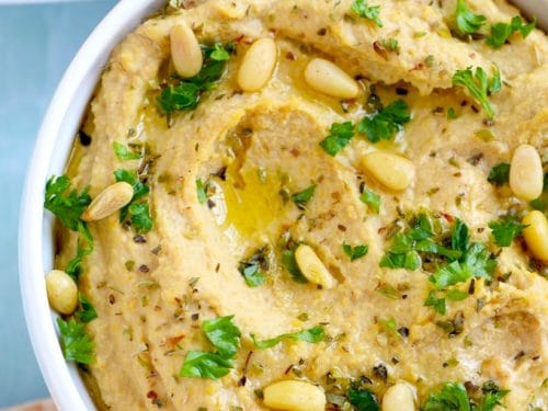 closeup of roasted garlic hummus in a white bowl with pine nuts and parsley