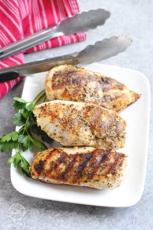 Grilled chicken breasts on a white plate with tongs and parsley