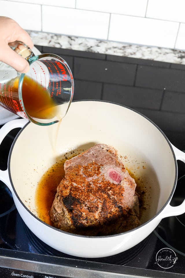 hand pouring beef broth into Dutch oven with seared beef inside