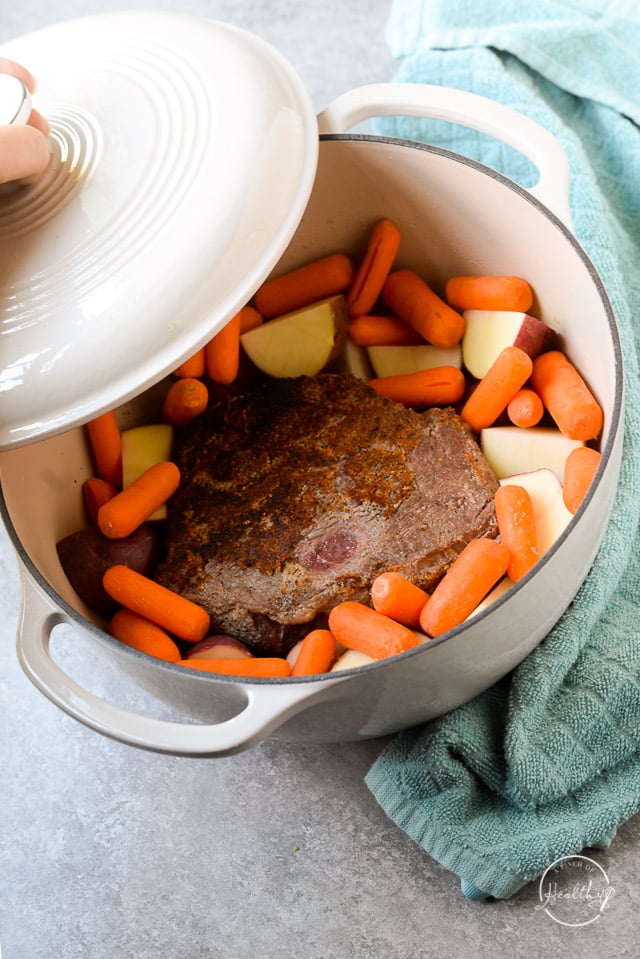 putting lid on white enameled Dutch oven with pot roast, potatoes and carrots inside