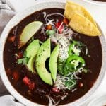 closeup of slow cooker black bean soup in a white bowl with avocado and tortilla chips