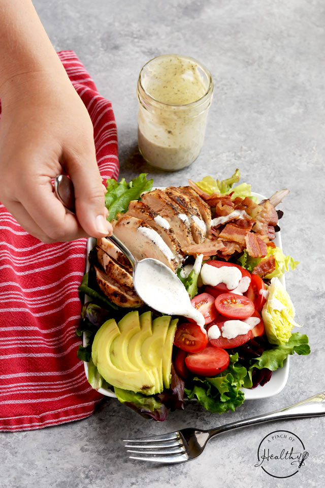 spooning ranch dressing onto a chicken BLT salad on a white plate