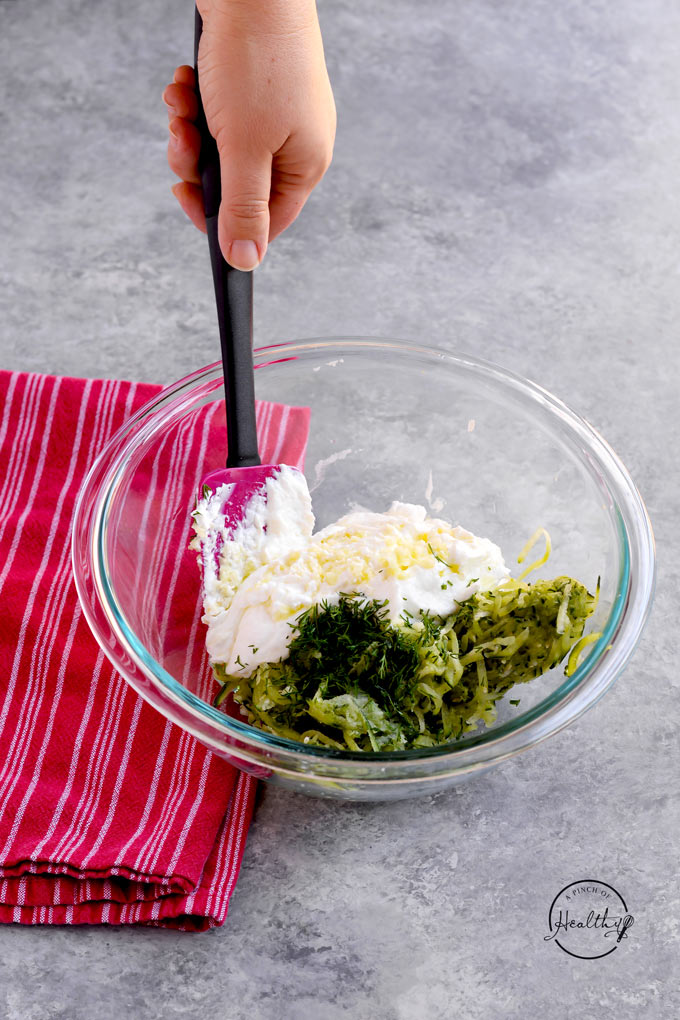 Stirring together individual ingredients for tzatziki in a clear bowl with a red towel