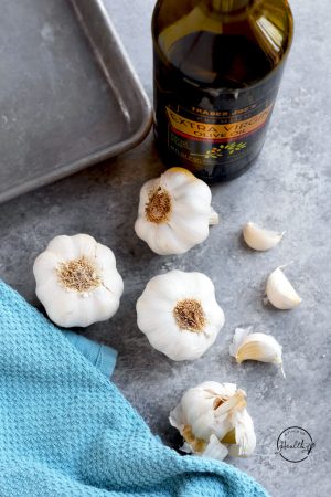 Garlic bulbs, plus a few cloves on a gray background with bottle of olive, baking sheet and blue towel