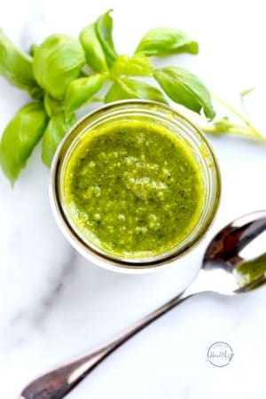 basil pesto in a mason jar on a marble surface with spoon and stems of fresh basil