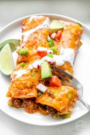 fork cutting into beef enchiladas on a white plate with slice of lime on the side