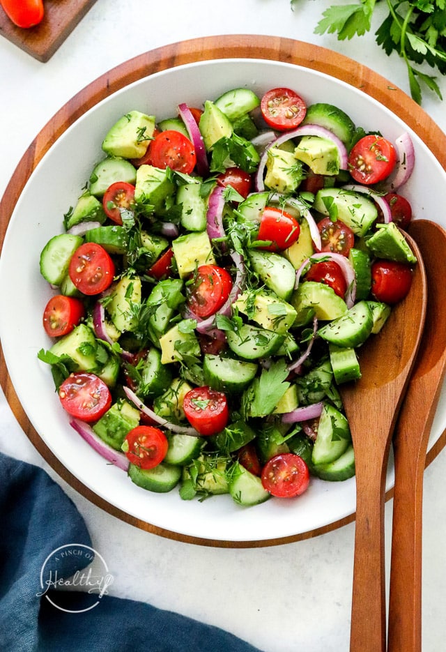 Tomato cucumber avocado salad with wood serving spoons in a white serving bowl