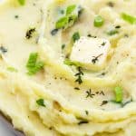 roasted garlic mashed potatoes in a serving bowl with spoon, herbs and a pat of butter