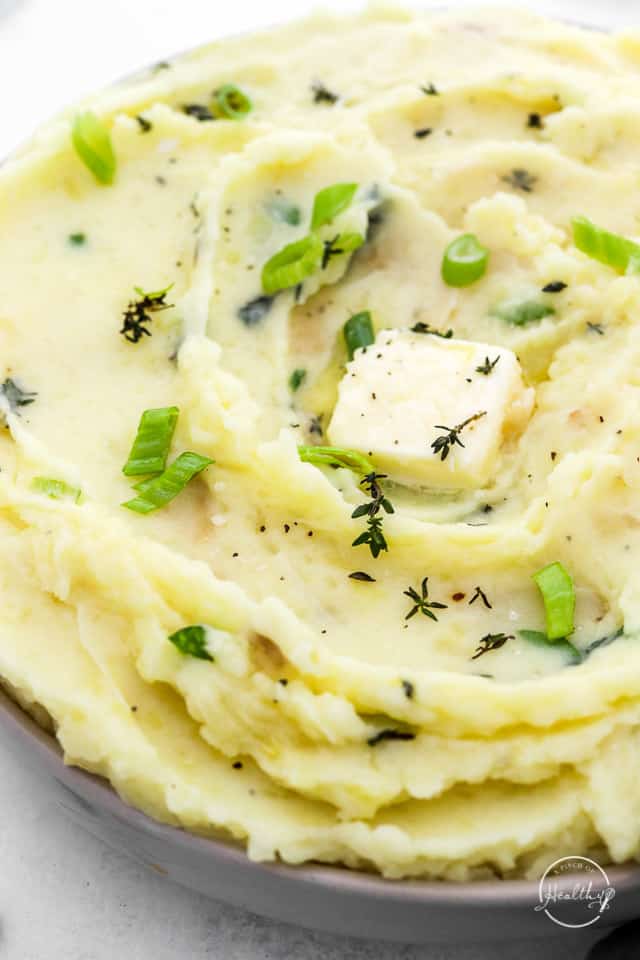 roasted garlic mashed potatoes in a serving bowl with spoon, herbs and a pat of butter