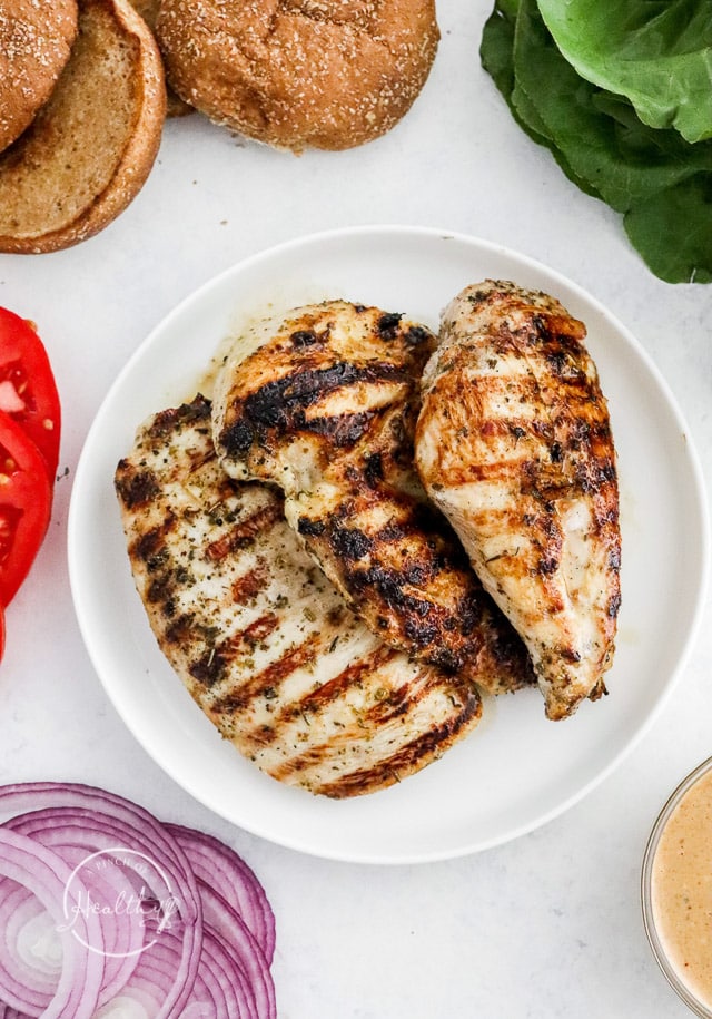 grilled chicken breasts on a white plate, surrounded by buns and veggies