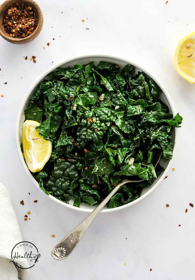 sautéed kale in a serving dish with lemon and spoon on white surface