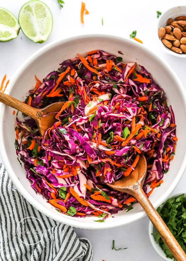 red cabbage slaw mixed in a white bowl with wooden serving utiensils
