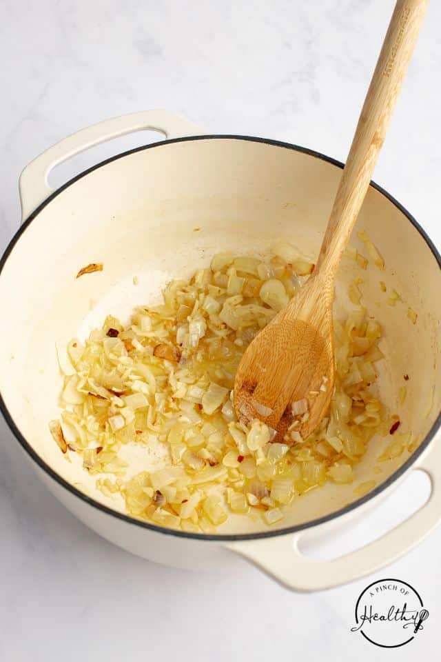 Onions, garlic, and red pepper in a Dutch oven cooking with olive oil and a wooden spoon in the pot