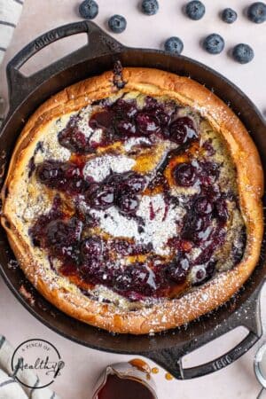 Blueberry Dutch Baby with maple syrup, blueberry compote, and powdered sugar in a cast iron pan.