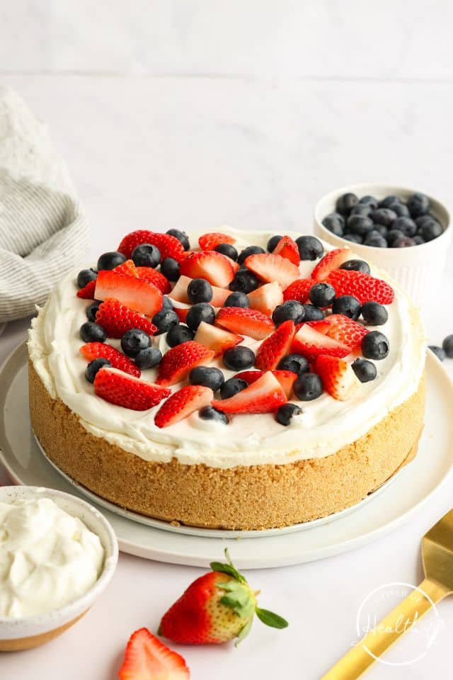 No bake cheesecake on a white table with berries on top and extra bowls of berries pictured.