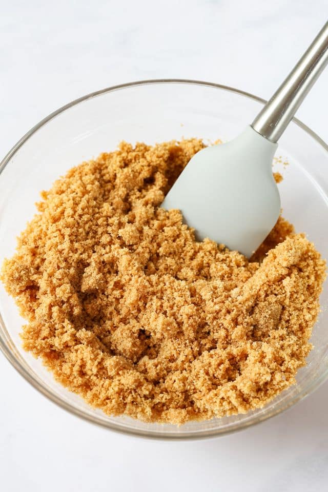 Ingredients for graham cracker crust in a glass bowl with a rubber scraper inside the bowl.