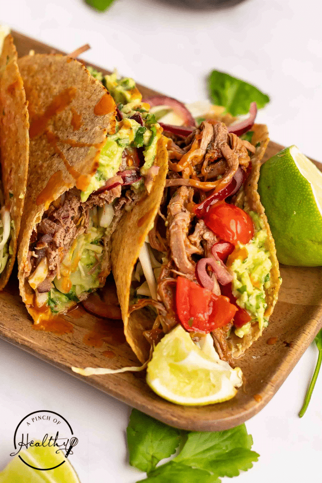 Brisket tacos on a wooden platter with lime wedges and tomatoes.