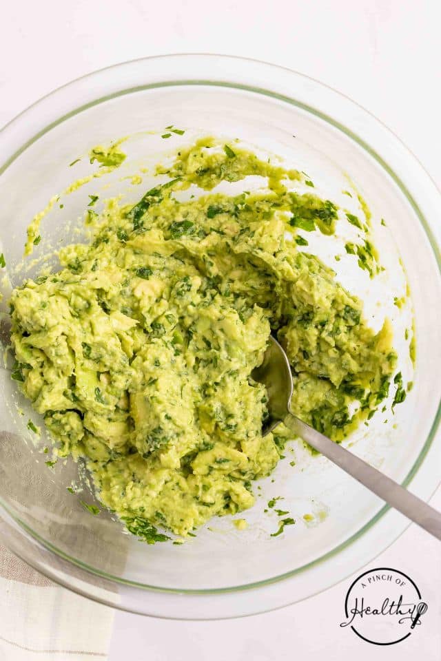 Guacamole ingredients mashed together in a glass bowl with a spoon inside the bowl.