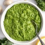 Kale pesto in a large white bowl with a silver spoon in the photo.