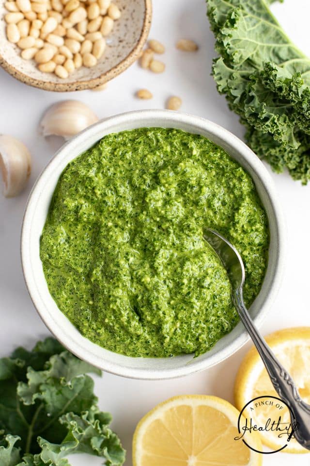 A large bowl of kale pesto on a white background with a spoon in the bowl and fresh kale and lemon slices on the side.