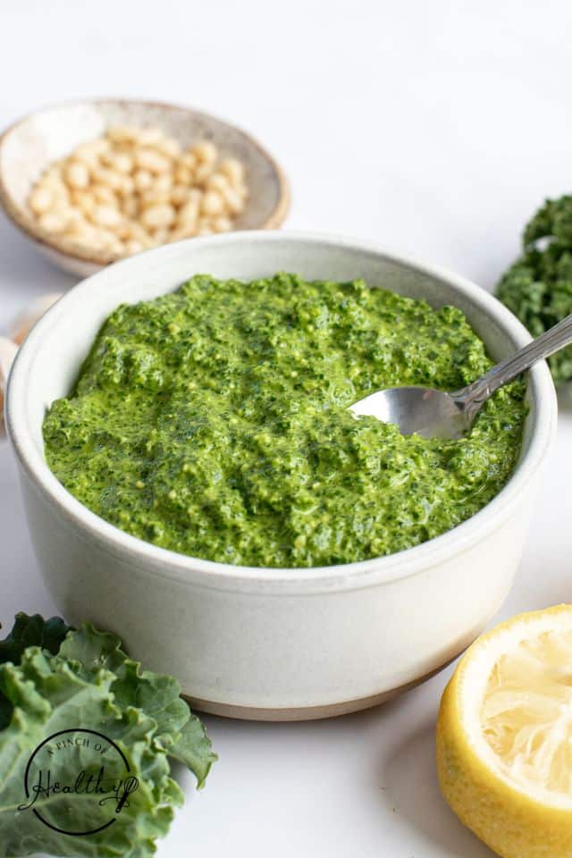 Kale pesto in a white bowl with a silver spoon in the bowl.