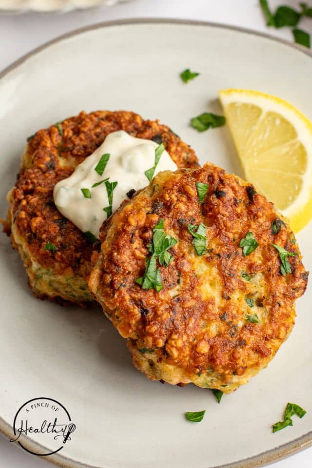 Two salmon cakes on a white plate with a slice of lemon and sauce on top.