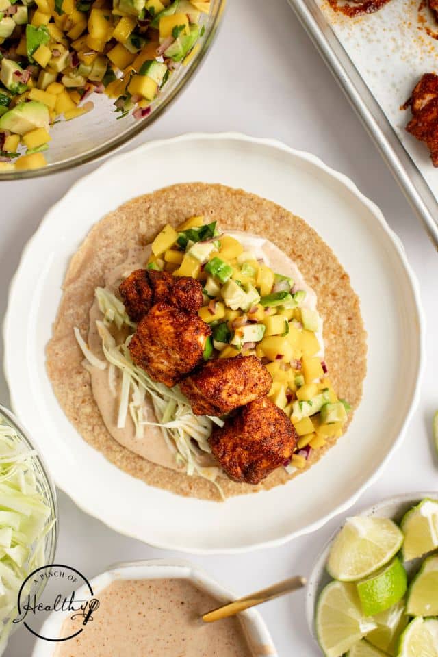 Assembled salmon taco on a white plate with the rest of the ingredients pictured.