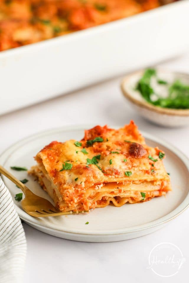 A slice of homemade lasagna on a white plate with a gold fork.