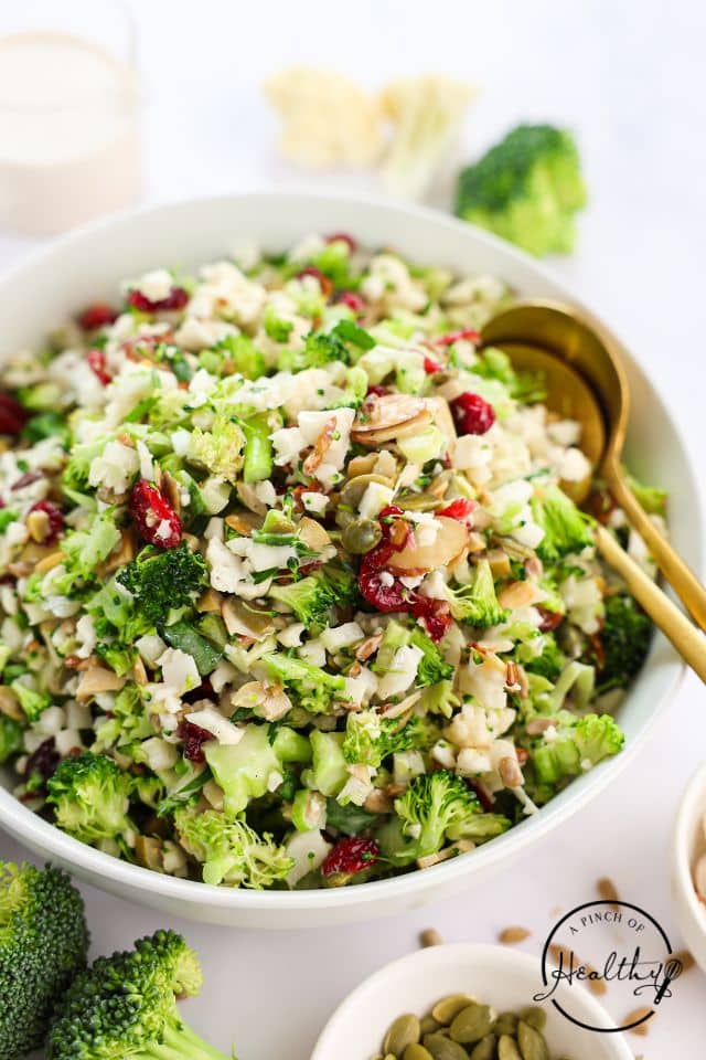 Broccoli cauliflower salad in a large white bowl with gold serving spoons.