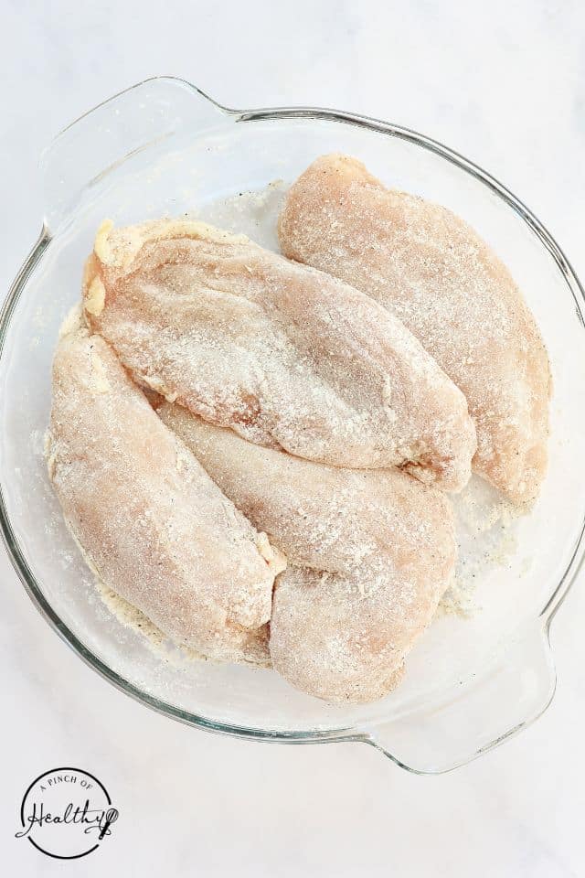 Chicken breasts dredged in flour and seasonings in a large glass bowl.
