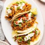 Shredded chicken tacos on a white oval platter with sour cream and tomatoes on top.