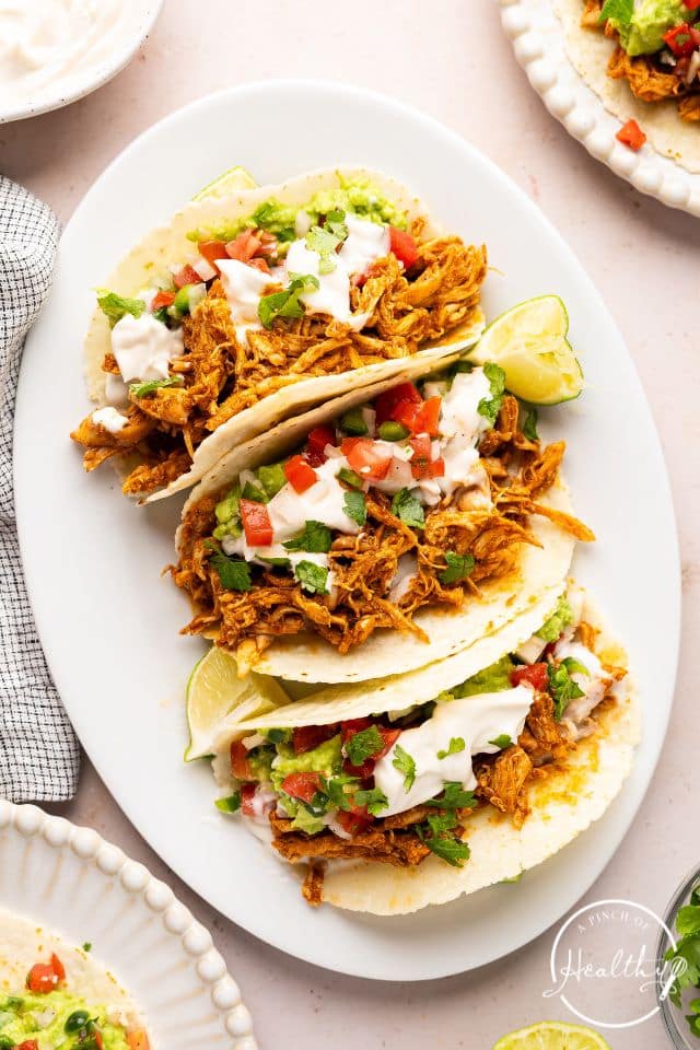 Shredded chicken tacos on a white oval platter with sour cream and tomatoes on top.
