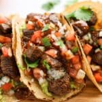 Steak tacos on a wooden cutting board with tomatoes.