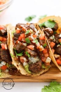 Steak tacos on a wooden cutting board with tomatoes.